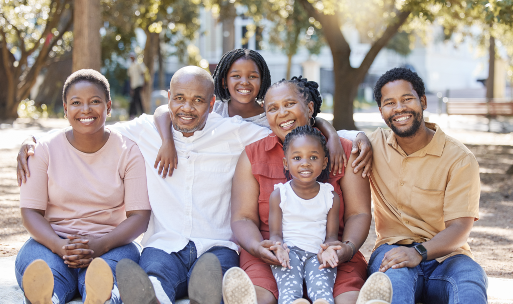 A joyful multigenerational family poses outdoors in a park. The group includes grandparents, parents, and two young children, all smiling warmly at the camera. The scene is set against a backdrop of trees and sunlight, creating a relaxed and happy atmosphere.